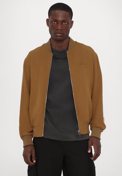 Veste bombardier marron avec fermeture éclair sur le devant, poignets et ourlet côtelés. Portée sur un t-shirt noir, présentant un détail de logo sur la poitrine gauche.