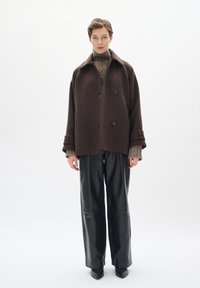 Chaqueta oversized de mezcla de lana marrón con grandes botones, combinada con un cuello de tortuga de canalé en color beige y pantalones de cuero negros de pierna ancha.