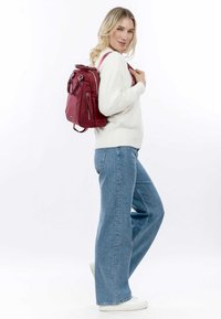 Rode leren rugzak met ritssluiting, twee schouderbanden en een handvat aan de bovenkant. Draag op de rug, gecombineerd met lichtblauwe flared jeans en een witte trui.