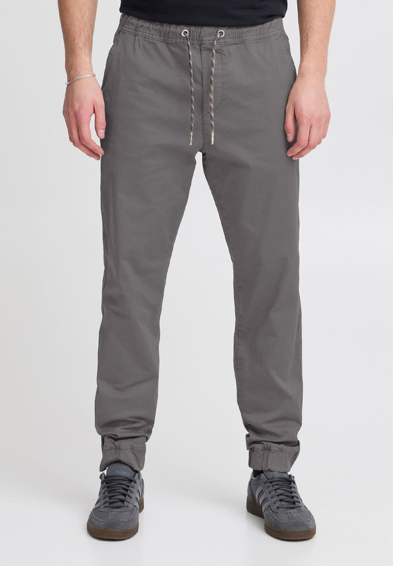 Mann trägt graue Jogginghose mit elastischem Bund und Kordelzug sowie graue Adidas-Sneaker, steht vor einem schlichten hellen Hintergrund.