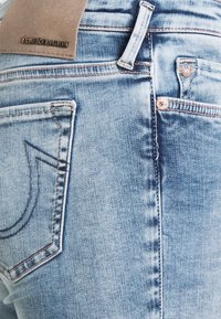 Jeans de mezclilla azul claro con una textura desgastada, que cuentan con un parche de cuero marrón, costuras en contraste y un diseño característico en el bolsillo trasero.