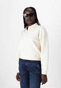 Felpa polo color crema con colletto e quattro bottoni, con logo ricamato sul petto. Abbinata a jeans blu scuro.