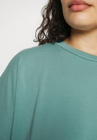 T-shirt de algodão em tom teal com decote redondo, mangas curtas e uma textura suave, apresentando detalhes de costura mínimos na gola.