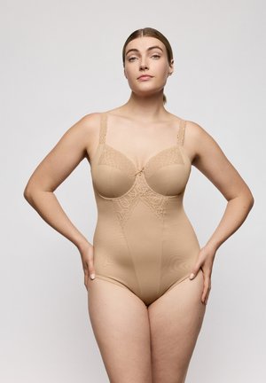 Vrouw met handen in de zij, gekleed in een beige body met kanten randjes, staand tegen een effen lichtgrijze achtergrond.