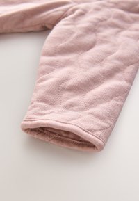 Quiltet lyserød stofærme med blød tekstur, med en rullet kant og subtilt mønster, placeret mod en neutral baggrund.