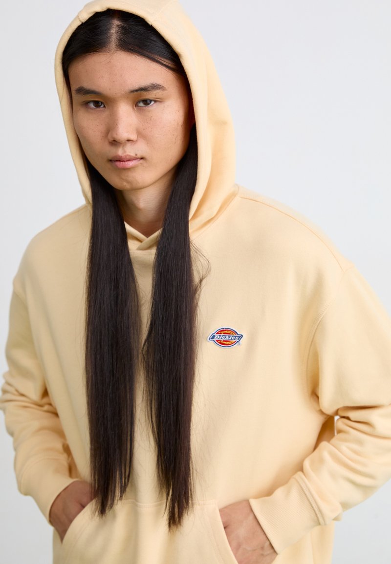 Sweatshirt à capuche jaune clair avec une poche avant, arborant un logo brodé circulaire. Fabriqué en tissu doux avec des poignets côtelés.