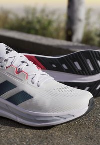 Baskets de sport blanches présentant une surface texturée, des rayures bleues, des accents rouges et une semelle extérieure noire avec des découpes pour une meilleure traction.