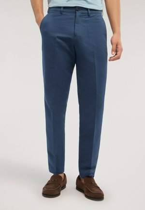 Homme portant un pantalon habillé bleu ajusté et des mocassins en daim marron, debout avec une main dans la poche devant un fond uni.