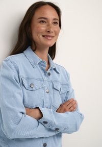 Gaiši zila strīpainā džinsu jaka ar priekšējiem kabatām un sudraba pogu aizdari; garas piedurknes ar pogām aizdarinātām, novietota izsistā veidā.