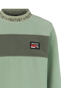 Šviesiai žalia pūkuota palaidinė, kurią puošia tamsiai žalia horizontali juosta ir juodas "PROTEST" logotipo pleistras su raudonais, baltais ir pilkais kalnų grafikos elementais.