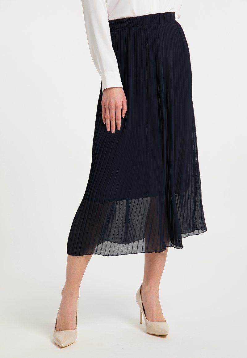 Femme portant une jupe midi plissée bleu marine, un haut blanc à manches longues et des chaussures à talons beiges, debout sur un sol blanc.