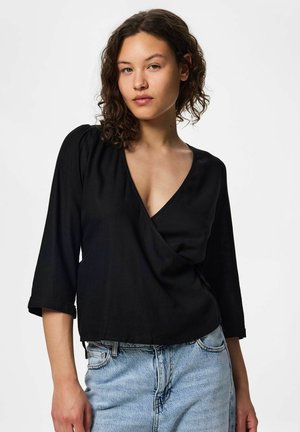 Tânără cu păr creț purtând un top negru în stil wrap cu decolteu în V și blugi albastru deschis pe un fundal simplu, deschis.