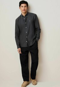 Camisa abotonada de color gris oscuro con un pequeño logo en el pecho, combinada con pantalones negros holgados y zapatos de lona beige. Fondo simple.