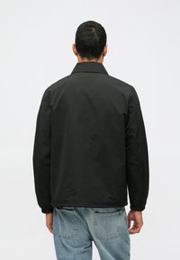 Μαύρο μπουφάν με χαλαρή εφαρμογή, με γιακά, ελαστικούς καρπούς και λεία υφή. Φορεμένο πάνω από μπλε τζιν παντελόνι.