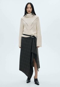 Maglione a collo alto a coste crema con dettagli a taglio, abbinato a una gonna avvolgente asimmetrica a quadri scuri e mule nere con tacco.