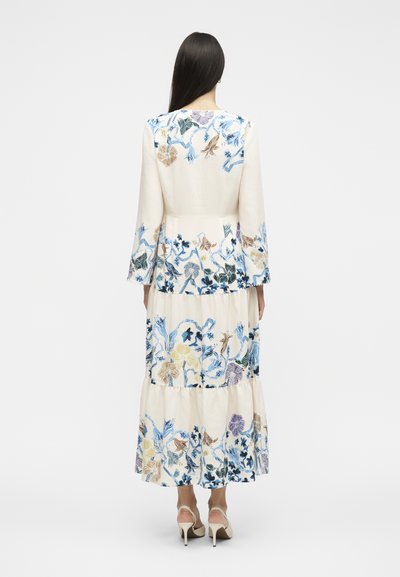Femme aux longs cheveux foncés portant une robe fleurie crème avec des fleurs bleues, violettes et beiges, debout de dos, portant des talons beiges.