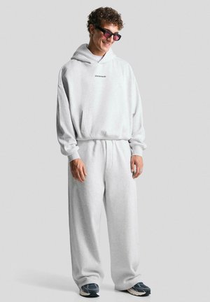 Mlad muž se smije, nosi svijetlosivu oversized duksericu i široke trenirke iste boje uz tamne tenisice i naočale sa zrcalnim staklima.