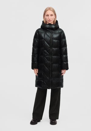 Femme portant un long manteau matelassé noir avec capuche, un pantalon foncé et des bottes noires, debout devant un fond clair uni.