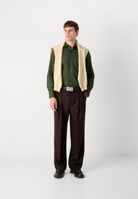 Chemise verte à manches longues, pull beige drapé sur les épaules, pantalon plissé marron, chaussures noires, boucle de ceinture argentée avec le logo "HUGO".