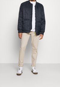 Přikrývková námořnická bunda bomber s žebrovanými manžetami, zipem vpředu a texturovaným povrchem, nošená přes bílou košili a spolu s béžovými kalhotami.