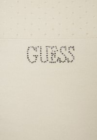 Krémová ribovaná látka s malými perforacemi, na které je slovo "GUESS" ve stříbrných kamíncích.