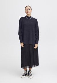Schwarzer gerippter Pullover, lockere Passform, hoher Kragen. Kombiniert mit einem schwarzen Faltenrock und schwarzen Hi-Top-Sneakern. Einfaches, elegantes Design.
