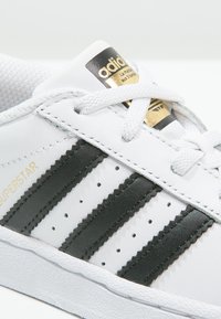 Sneaker bianca con tre strisce nere, dettagli perforati, suola in gomma testurizzata, logo dorato in rilievo sulla linguetta, chiusura con lacci.