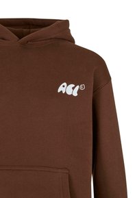 Sudadera con capucha marrón con un bolsillo tipo canguro en la parte frontal. Presenta un logotipo gráfico blanco en el lado izquierdo del pecho. Fabricada con un tejido suave y texturizado.
