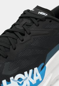 Black athletic shoe featuring textured mesh upper, black laces, white and blue branding on the side, and a chunky white sole.