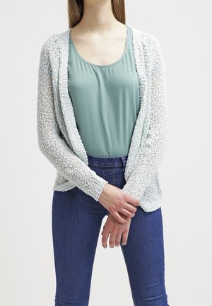 Femme portant un haut sans manches bleu clair, un cardigan blanc à mailles ouvertes et un jean taille haute bleu, debout les mains jointes devant elle.