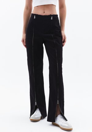 Zwarte broek met een hoge taille, voorzien van ritsen aan de voorkant, brede pijpen en onafgebouwde randen in de taille. Gestyled met witte sneakers.