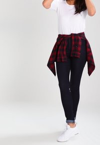 Camiseta blanca de manga corta combinada con jeans ajustados azul oscuro, camisa de cuadros roja y negra atada a la cintura y zapatillas blancas.