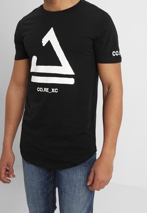 T-shirt noir avec un motif abstrait blanc de triangles et de lignes, texte « CO.RE_XC » sur le devant et la manche, porté avec un jean bleu par une personne debout.