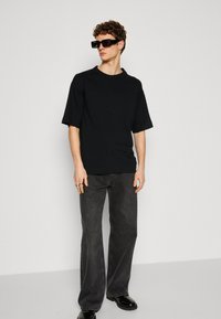 T-shirt nera oversize con maniche corte, abbinata a jeans a zampa d'elefante grigio scuro e scarpe nere. Modella che indossa occhiali da sole rettangolari.