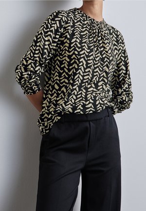 Persoon die een zwart-beige blouse met lange mouwen en patroon draagt, ingestopt in een zwarte broek met hoge taille, staand met handen achter de rug.