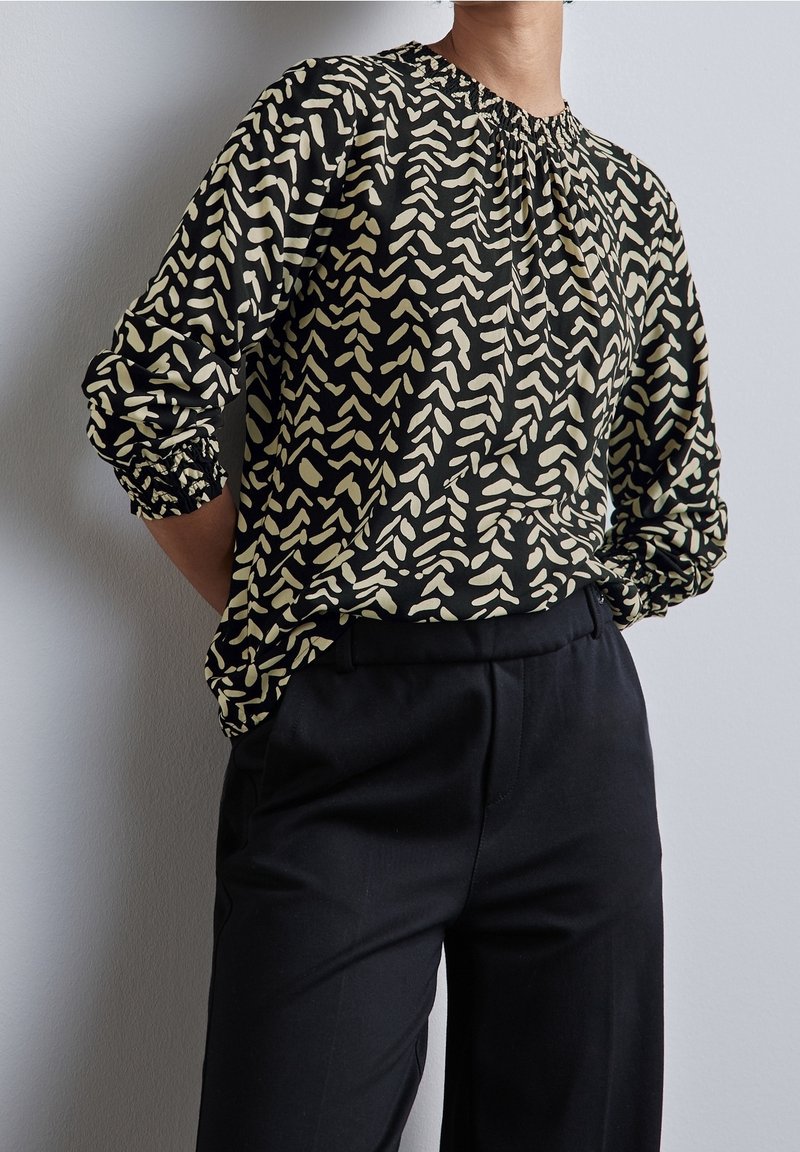 Personne portant un chemisier à manches longues noir et beige à motifs, rentré dans un pantalon noir taille haute, debout les mains derrière le dos.
