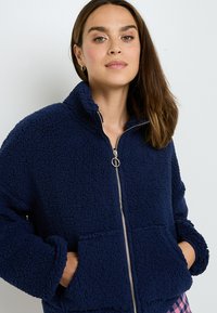 Donna in giacca di pile strutturato blu navy con zip e mani in tasca su sfondo neutro.