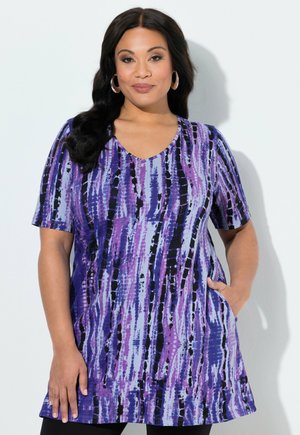 Kobieta o długich ciemnych włosach, nosząca kolczyki koła oraz tunikę z krótkim rękawem i dekoltem w serek w fioletowo-czarno-niebieski wzór, z ręką w kieszeni.