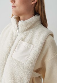 Ivoren fleece vest met hoge kraag, ritsluiting en een vierkante satijnen zak. Getextureerd materiaal met een zachte, pluizige oppervlak.