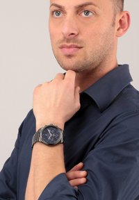 Schwarze Metallarmbanduhr mit einem runden blauen Zifferblatt, silbernen Akzenten und einem Gliederarmband. Der Träger ist in einem dunkelblauen Hemd gekleidet.