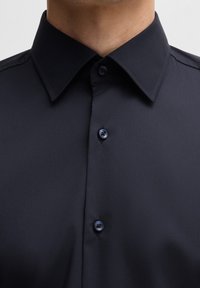 Camicia blu navy con bottoni e colletto standard, trama liscia e quattro bottoni abbinati sul davanti. Vista ravvicinata della parte superiore.