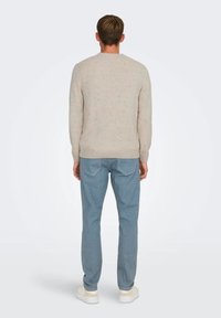 Suéter de punto beige con manchas, cuello redondo y puños acanalados, combinado con jeans de color azul claro y zapatillas blancas, visto desde la parte de atrás.