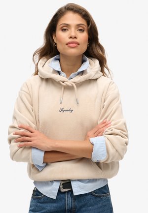 Sudadera de pana beige con cordón, logo bordado, superpuesta sobre una camisa de botón azul clara. Modelo con los brazos cruzados frente a un fondo neutro.