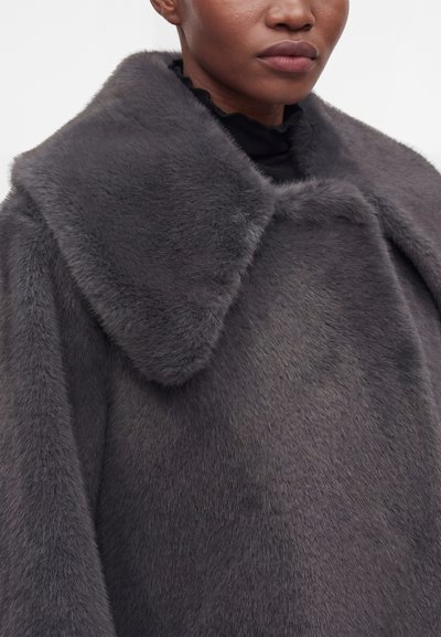 Gros plan d'une personne portant un manteau en fausse fourrure gris foncé avec un grand col et un col roulé noir en dessous.