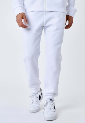Persona che indossa pantaloni jogger bianchi e sneaker Nike Air bianche con swoosh nero su uno sfondo bianco uniforme.