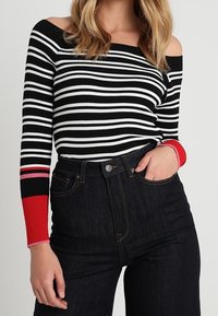 Zwart-wit gestreepte ribbel top met off-the-shoulder halslijn en rode accentmouwen. Gepaarde met hoog-getailleerde donkere spijkerbroeken.