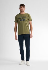 Olivengrøn bomulds-T-shirt med mørkeblå tekst, der står "NASCAR CUP DAYTONA PETROL IND. EST. 1989." Kombinationen er med marineblå bukser og hvide sneakers.
