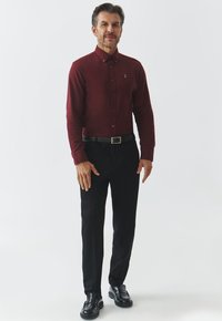 Maronfarvet skjorte med knaplukning og krave, sorte bukser og skinnende sorte loafers. Simpelt design med et lille logo på venstre bryst.