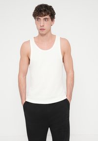 Λευκό ριμπ τοπ με φαρδιές τιράντες· συνδυασμένο με μαύρο παντελόνι με υφή και πλαϊνές τσέπες. Το μοντέλο έχει τα χέρια στις τσέπες.