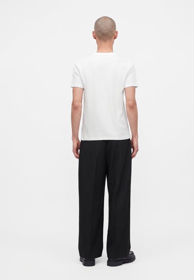 Homme debout de dos, portant un t-shirt blanc uni à manches courtes, un pantalon noir large et des chaussures noires sur fond blanc.